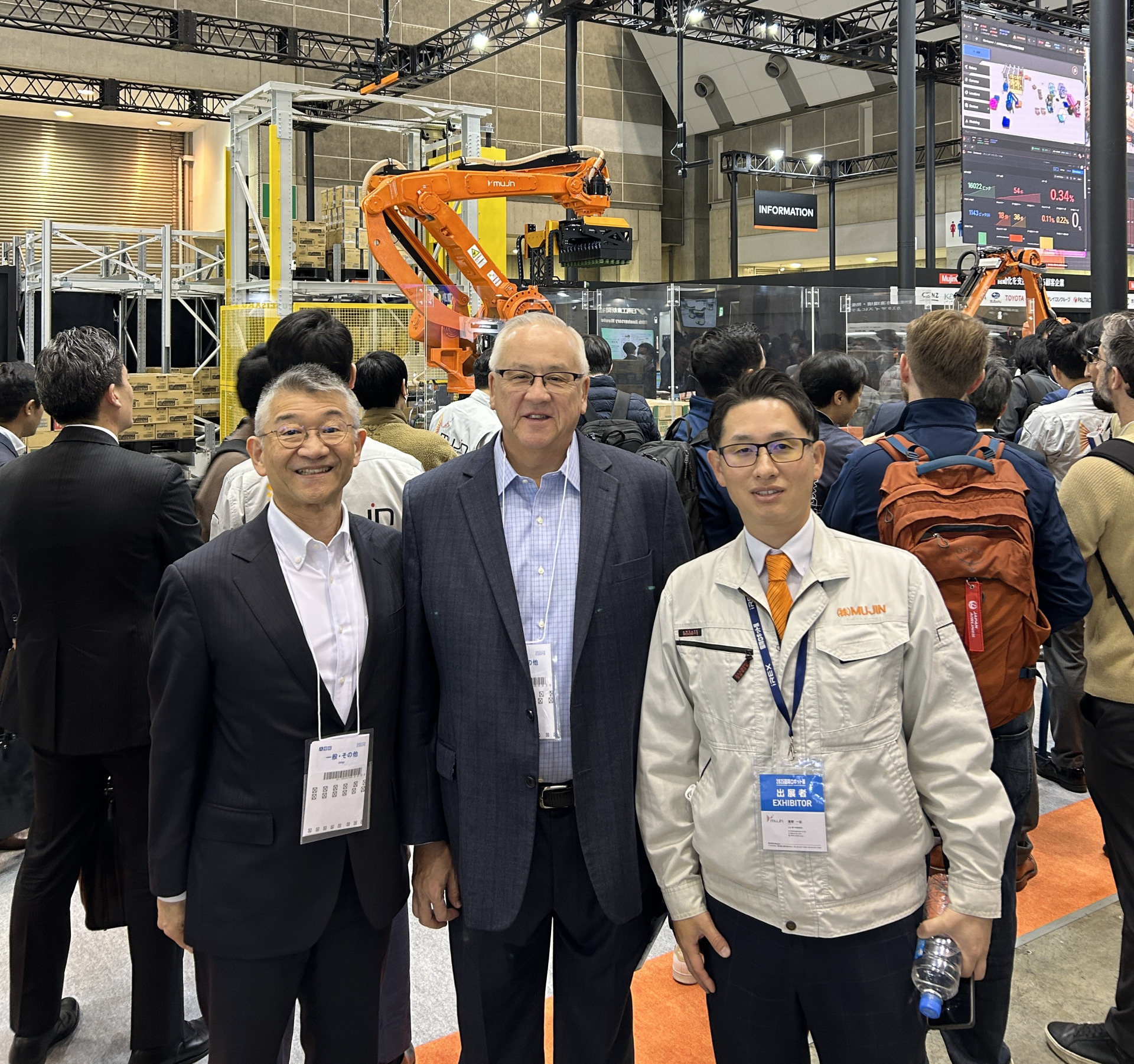 Two men wearing suit jackets and dress shirts and a third man wearing a jacket with "Mujin" embroidered on it smile and pose for a photo together at the 2025 International Robot Exhibition in Tokyo.