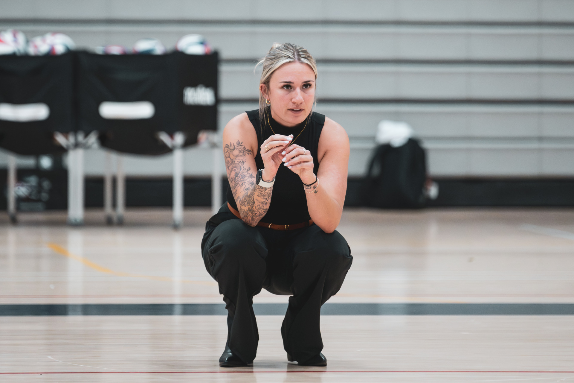 A blonde woman with her hair up, wearing a black tanktop and pants squats down on a gym floor with volleyballs in the distance.