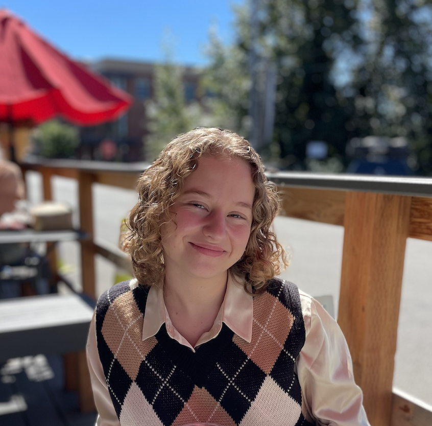 A woman with curly dirty blonde hair wears a checkered vest and smiles while sitting on a patio outside with a blue sky and red umbrella above.