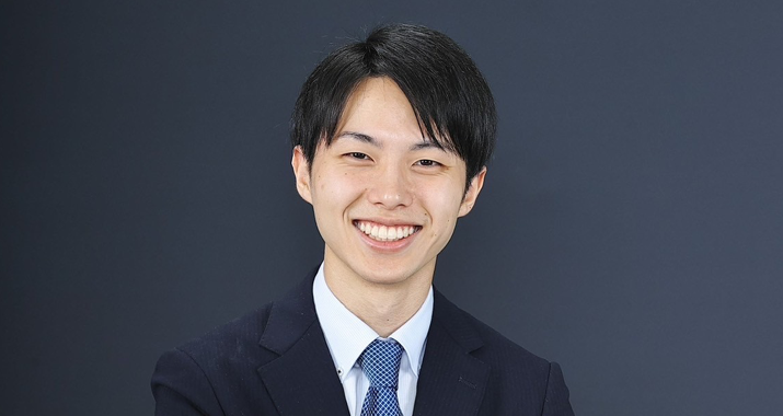 A man with dark brown hair wearing a dark suit with a lighter blue tie smiles.