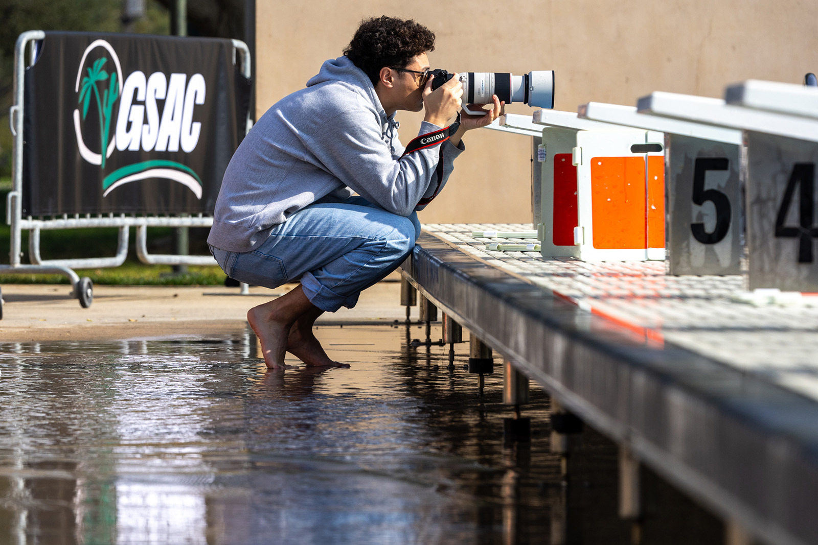 A barefoot photographer in a grey hoodie and jeans crouches on a wet pool deck, aiming a Canon camera with a large telephoto lens toward the starting blocks. In the background, a banner reads "GSAC."
