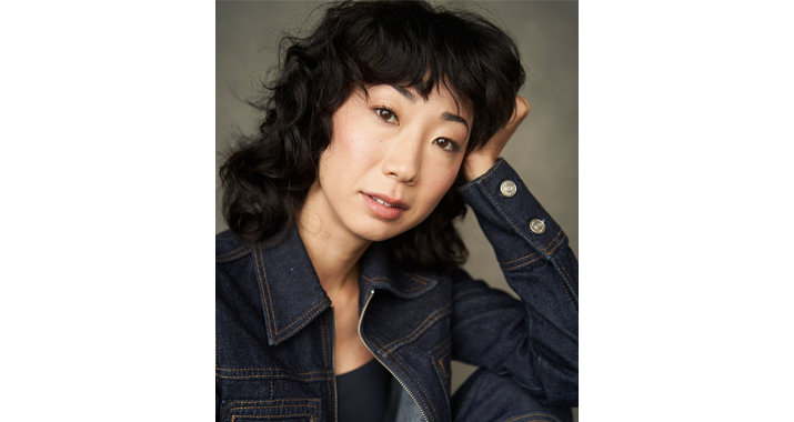A woman with curly dark brown hair smiles and tilts her head to the right. She is wearing a jean jacket and resting her head in her left hand.
