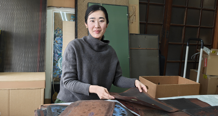 A woman with black, pulled-back hair, wears a grey turtleneck sweater and is working with a pile of Japanese textiles at a table. 