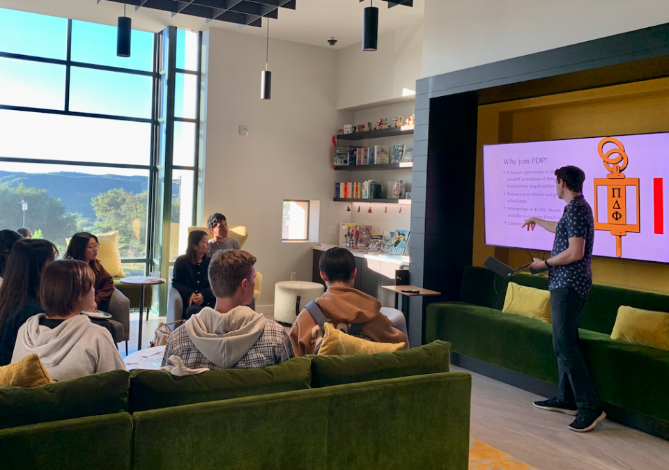 A man points to a tv screen while presenting to a group of students sitting on a green velvet couch.