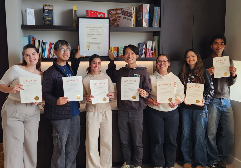 Seven students hold certificates and pose in front of a bookshelf. Two of the students are also displaying a framed certificate.