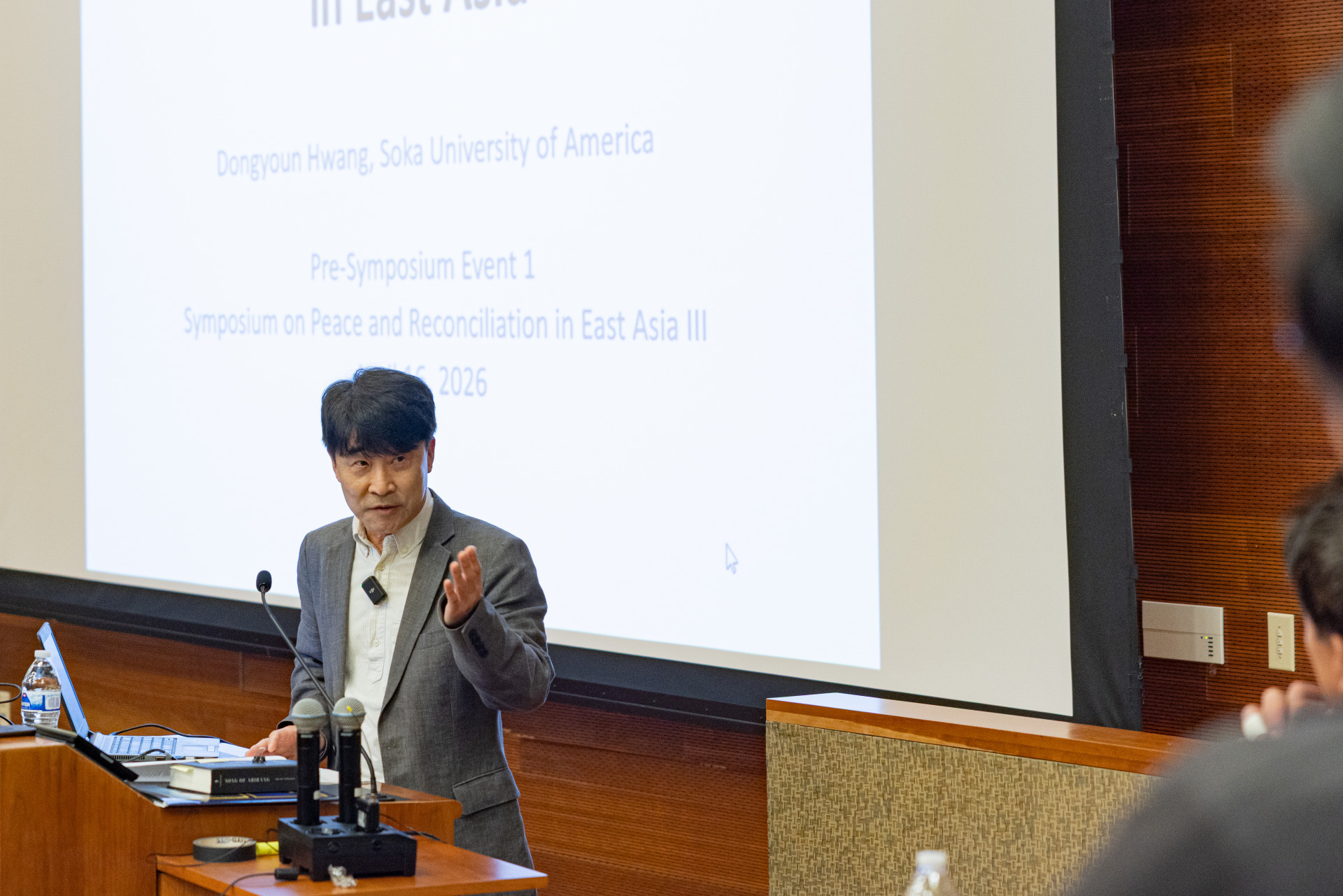 A man with short black hair is wearing a gray suit and gestures with his left hand as he stands in front of a projector screen while giving a presentation.