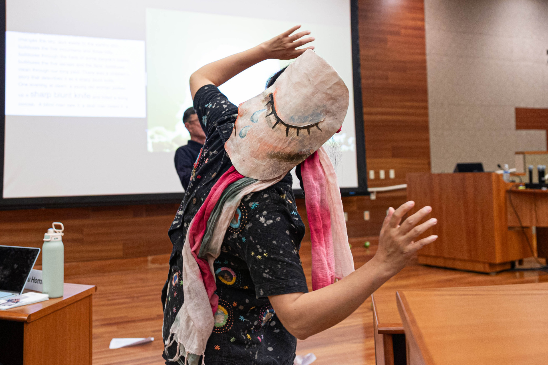 A person wearing a mask depicting a closed, crying eye on their head performs and holds both of their hands in the air.