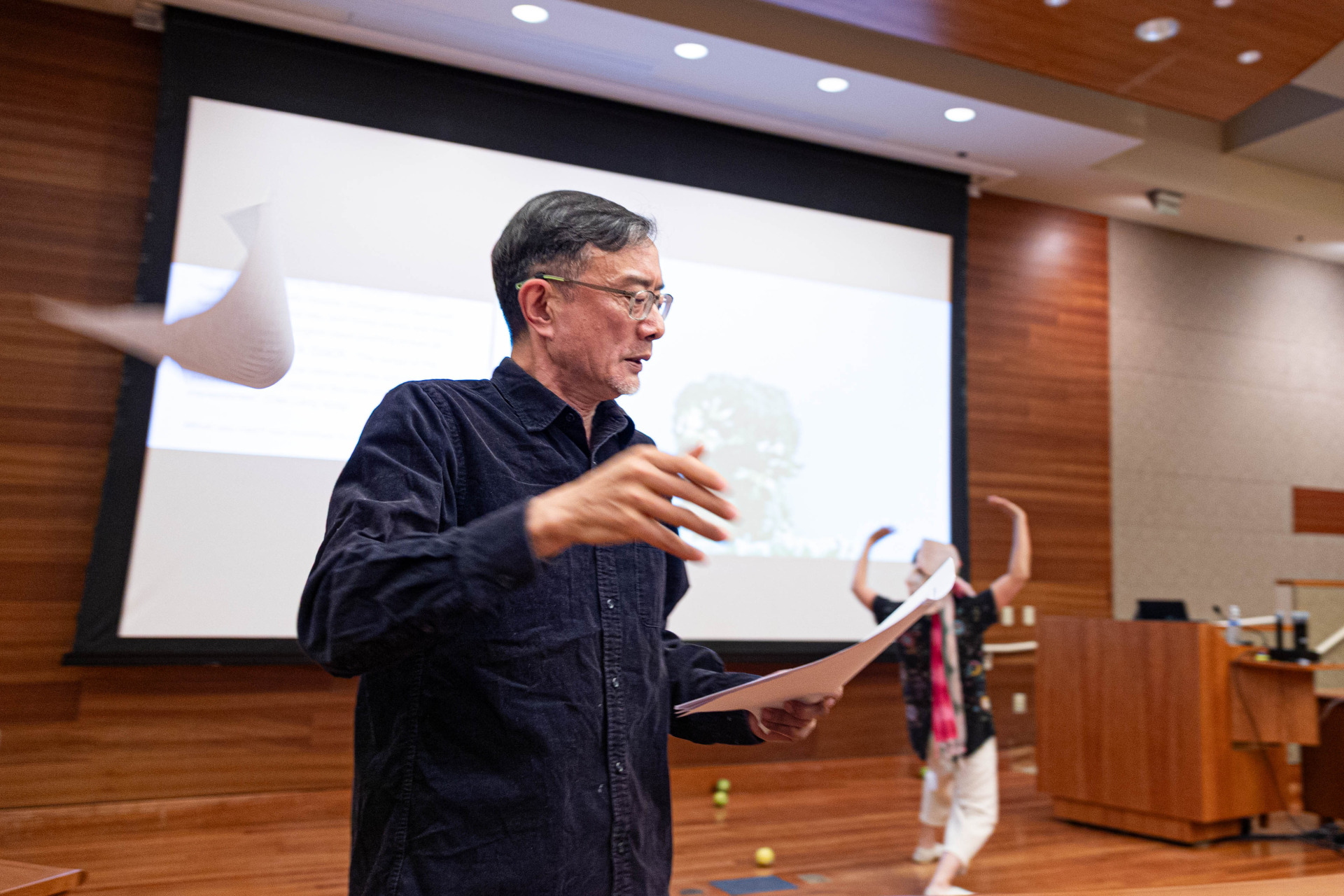 A man with short black hair and glasses wearing a navy button up holds a stack of white papers and throws one into the air during a theatrical performance inside a classroom auditorium.
