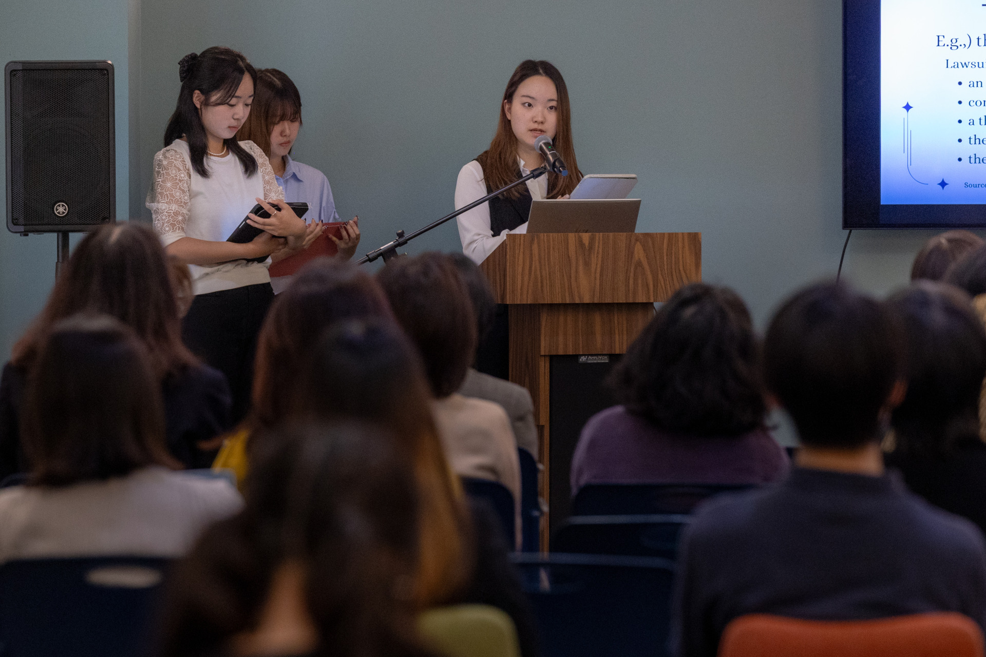 Three people wearing professional attire stand at the front of the room. One is speaking into a microphone at a wooden lectern to a seated audience. 