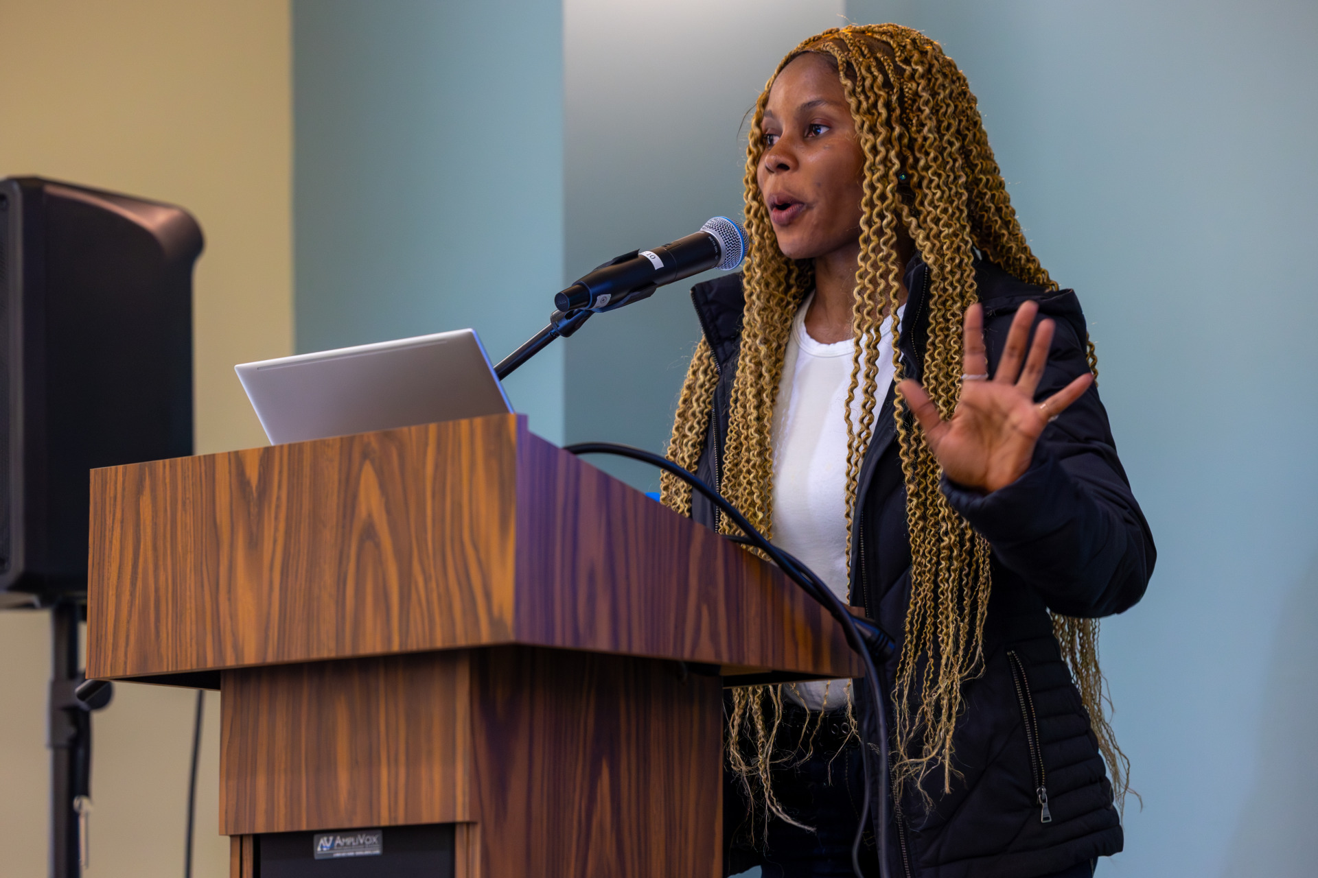 A woman with long curly blonde braids wearing a white t-shirt and blue jacket speaks into the microphone behind a wooden lectern.