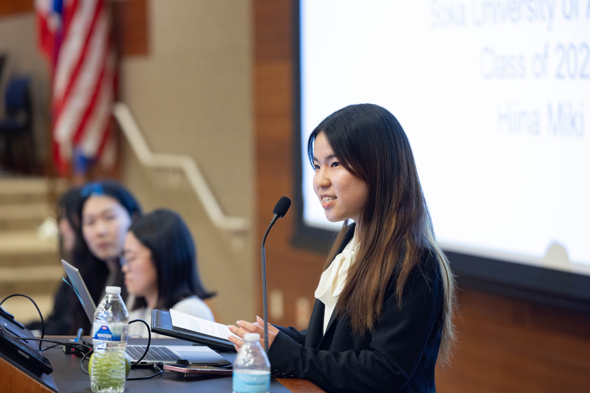 A woman with long brown hair wearing a dark suit jacket over a white shirt speaks into the microphone during a presentation.