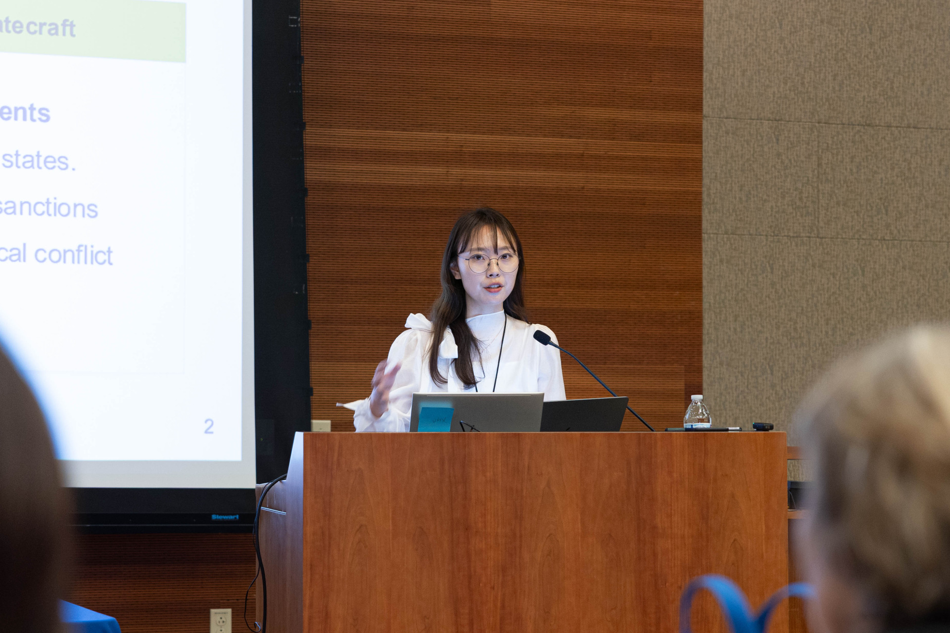A woman with long dark hair wearing a white blouse speaks into the microphone at a wooden lectern during a presentation.