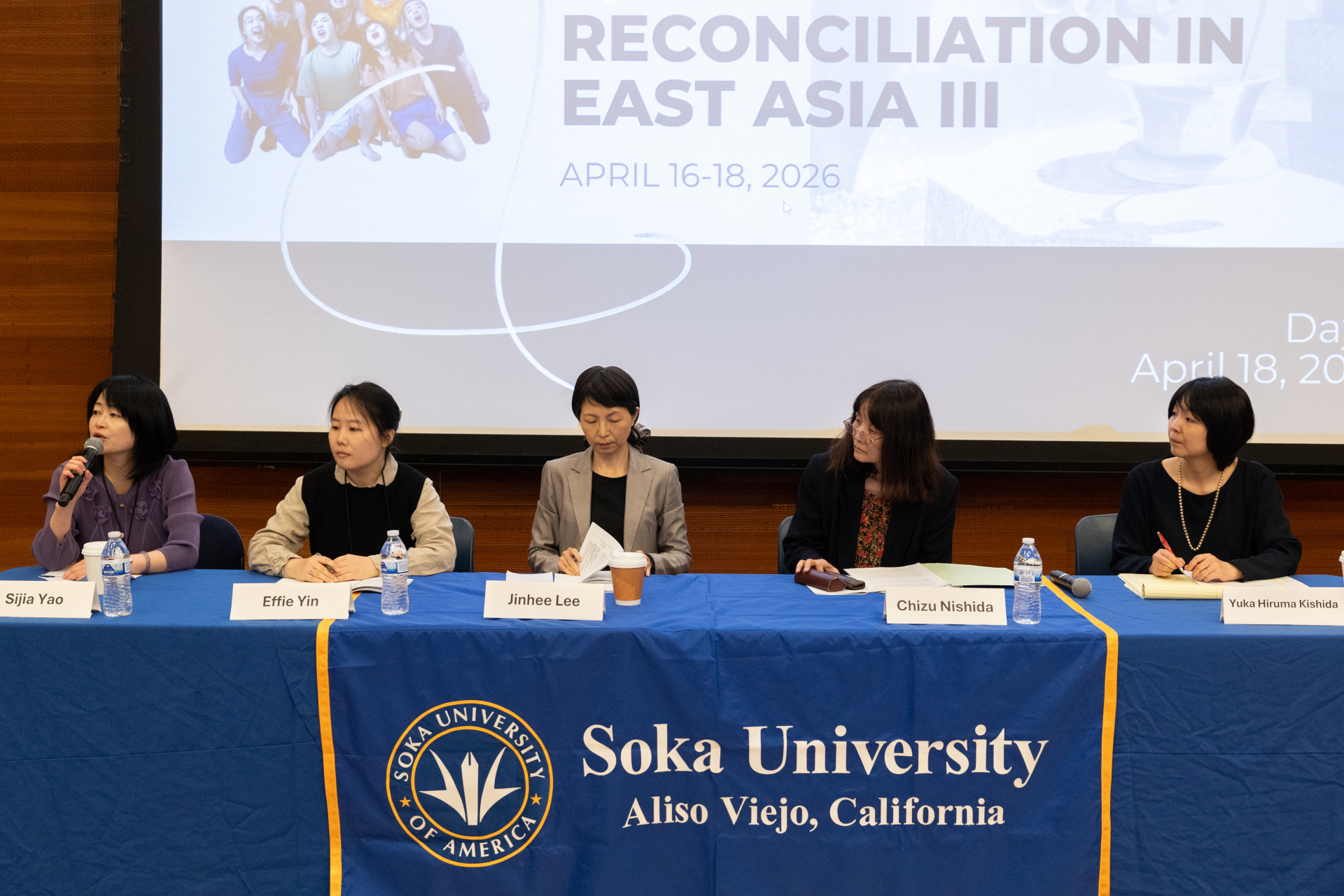 A panel of people sit behind a long table covered with a royal blue tablecloth that contains Soka's logo. They are sitting in front of a projector screen that has conference information on it.