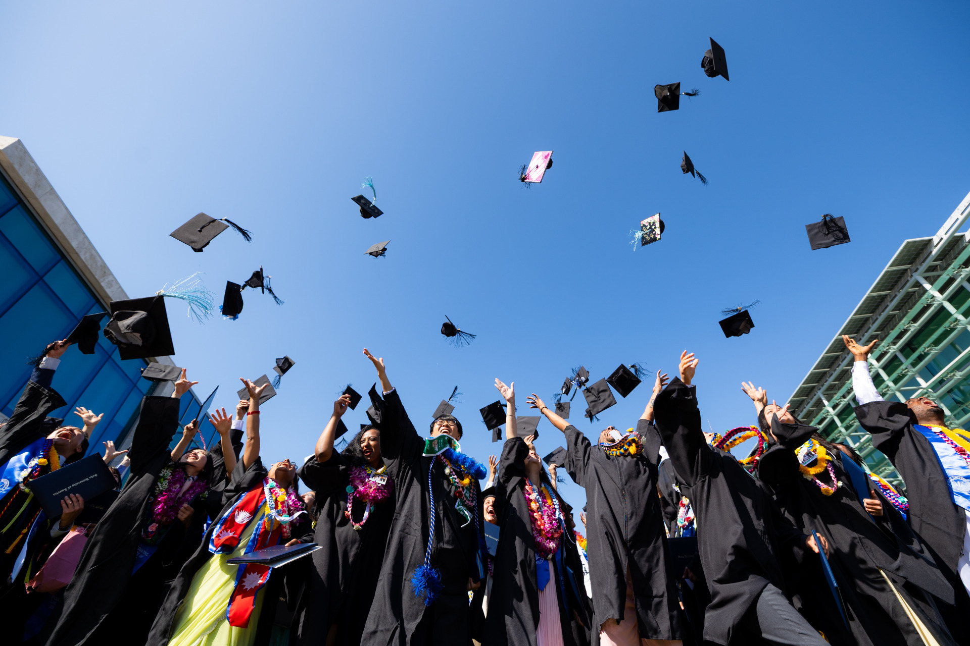 SUA Graduates wearing a mix of colorful clothes under their black graduation gowns throw their caps into the air in celebration.