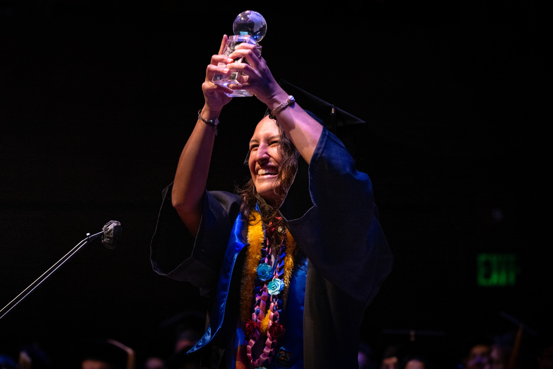 A woman with curly brown hair smiles and holds a clear globe statue award up in both arms. She is wearing a black graduation cap and gown with yellow and blue cords.