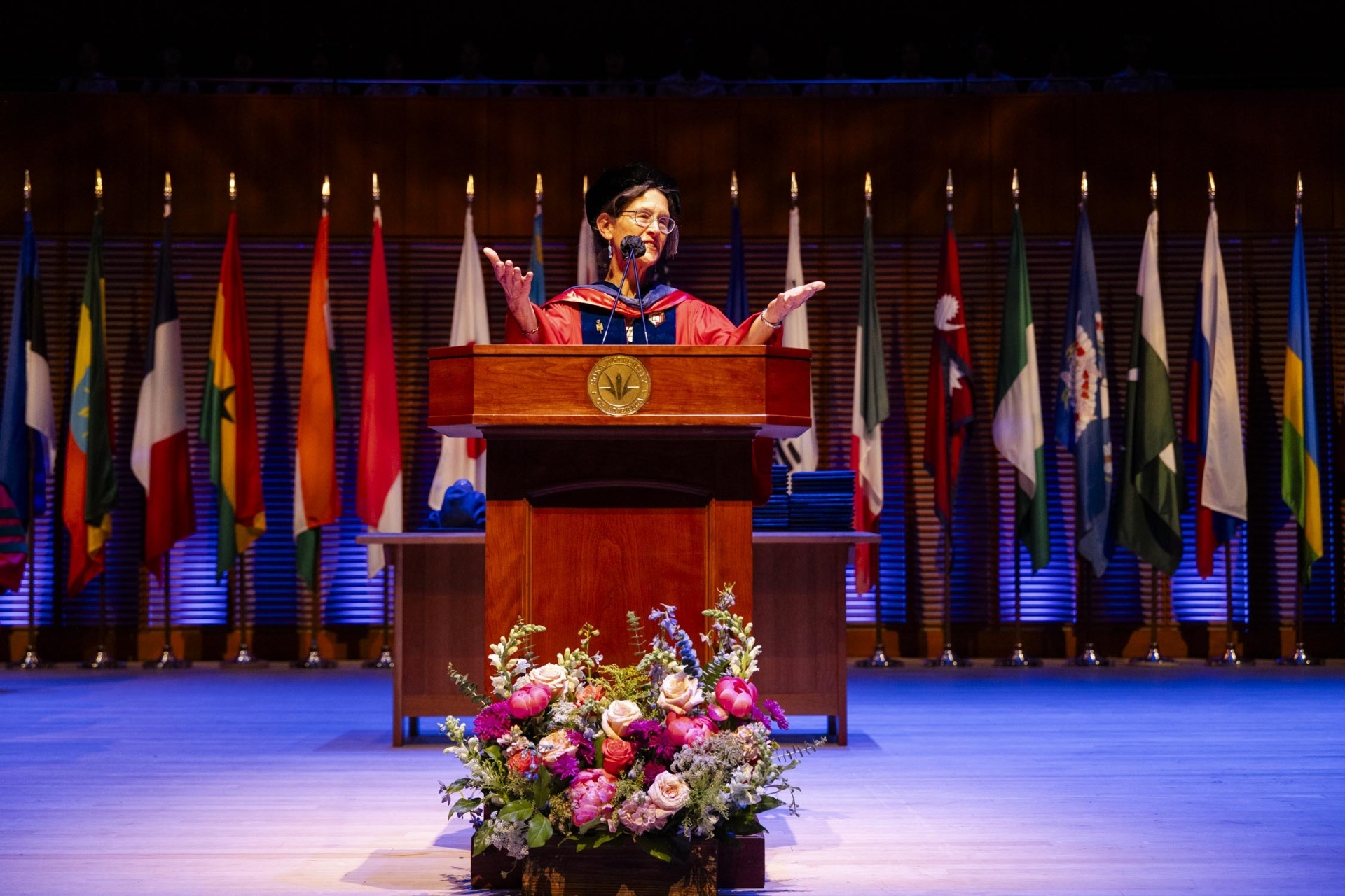 Dr. Sarah Ann Wider is wearing a red graduation rob and black cap and glasses. She holds her palms up as she is standing behind a wooden lectern with a black microphone on it. There is a vase of flowers at its base and numerous country flags lined up behind her.