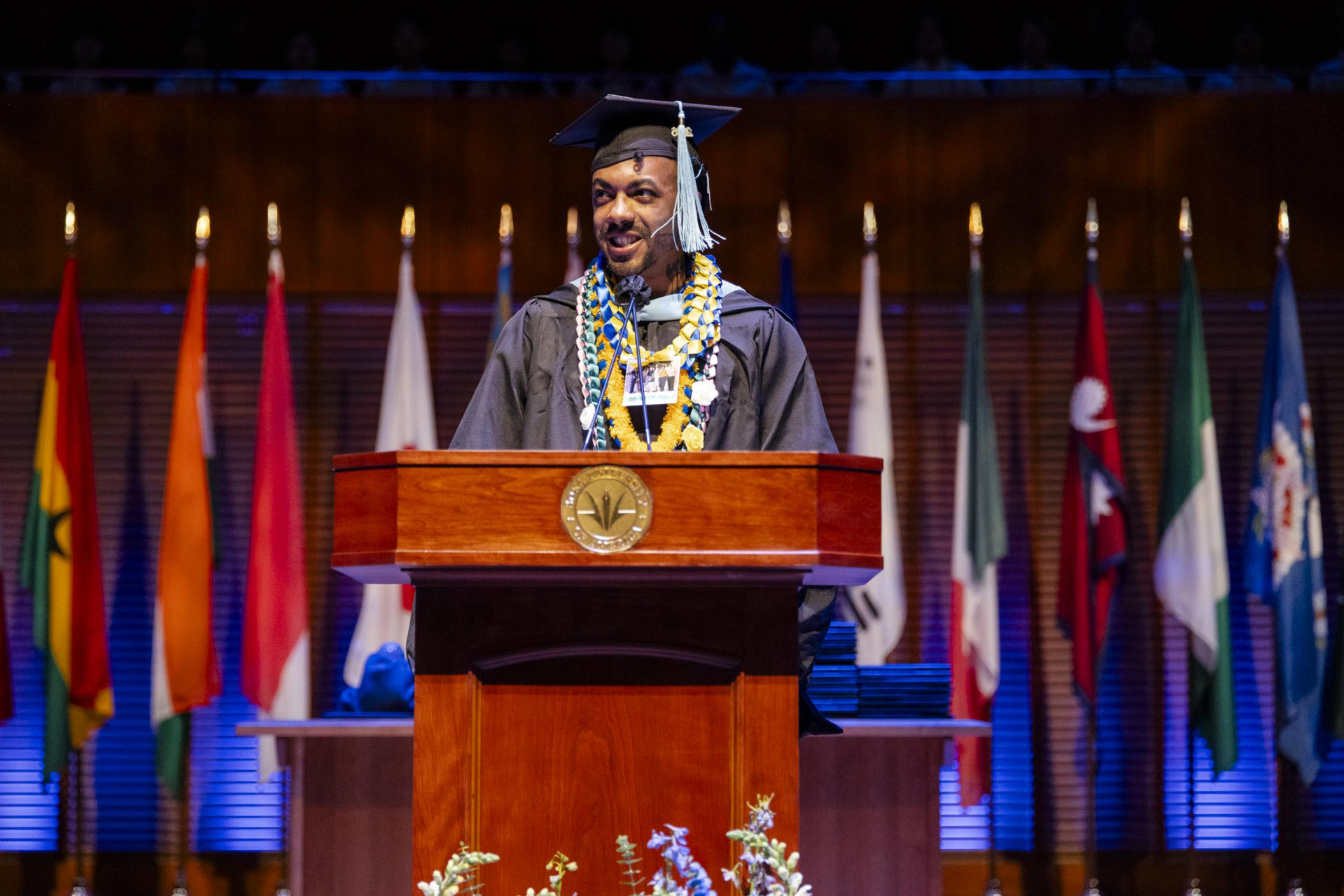 A graduating student wearing a black cap and gown with a light blue tassel speaks at a wooden lectern in front of a horizontal line of country flags.