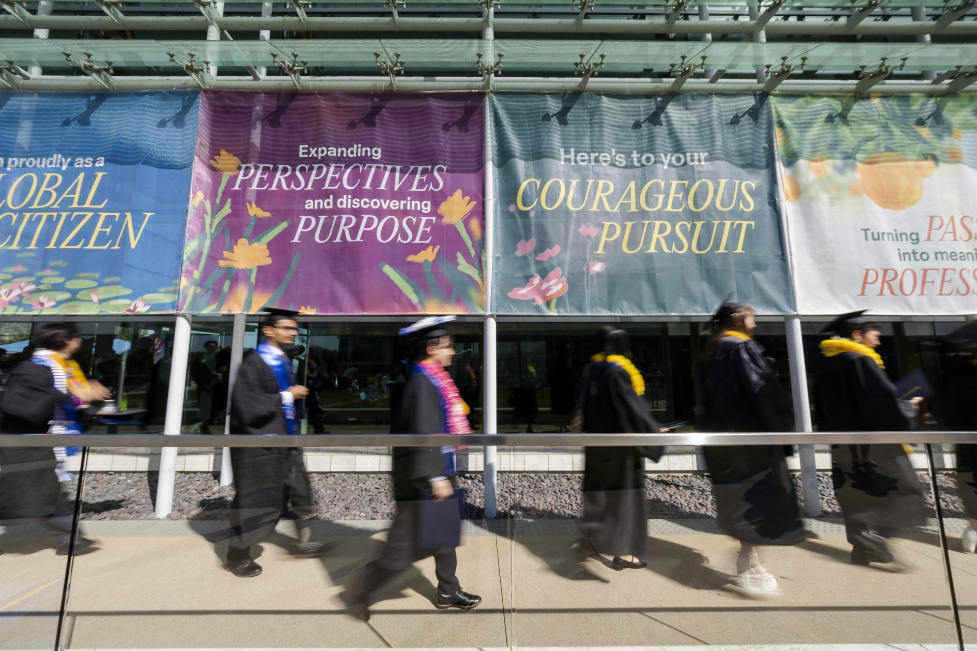 Graduates wearing black caps and gowns with yellow, pink, and blue cords and leis, walk past colorful marketing signs on the outside of the Performing Arts Center.