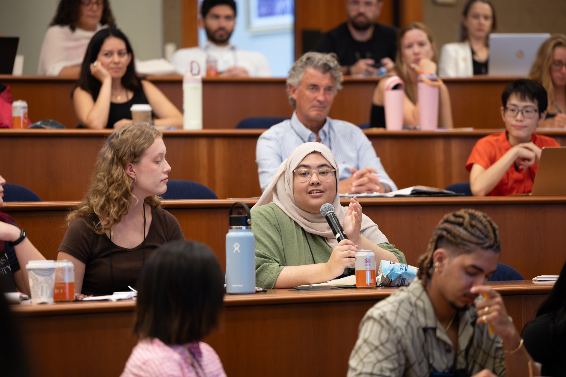 A woman wearing a tan headscarf and green shirt speaks into a microphone as people sitting around her listen on.