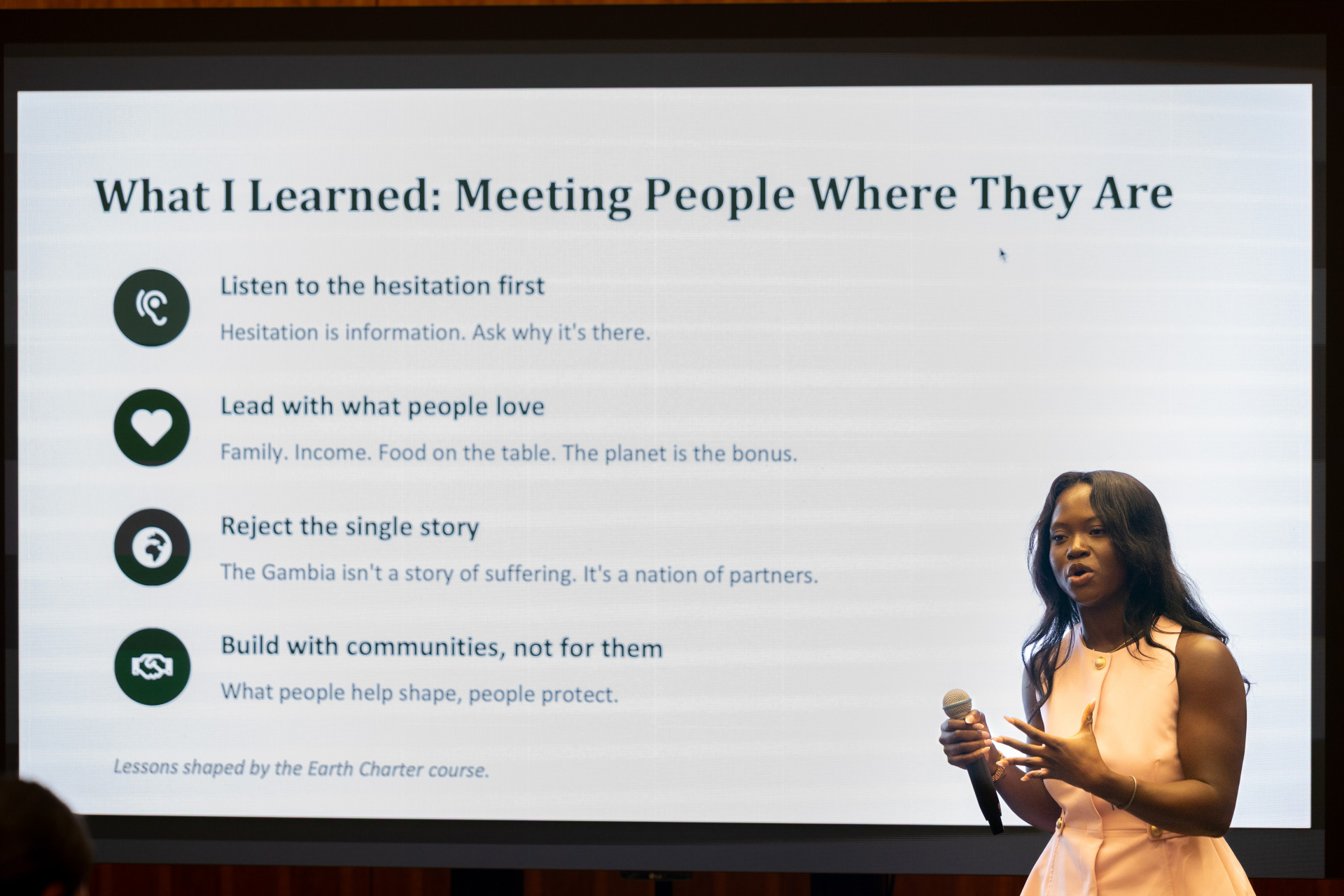 Awa Drummeh, a woman with black hair wearing a peach colored dress holds a microphone and stands in front of a projector screen while she gives a presentation.