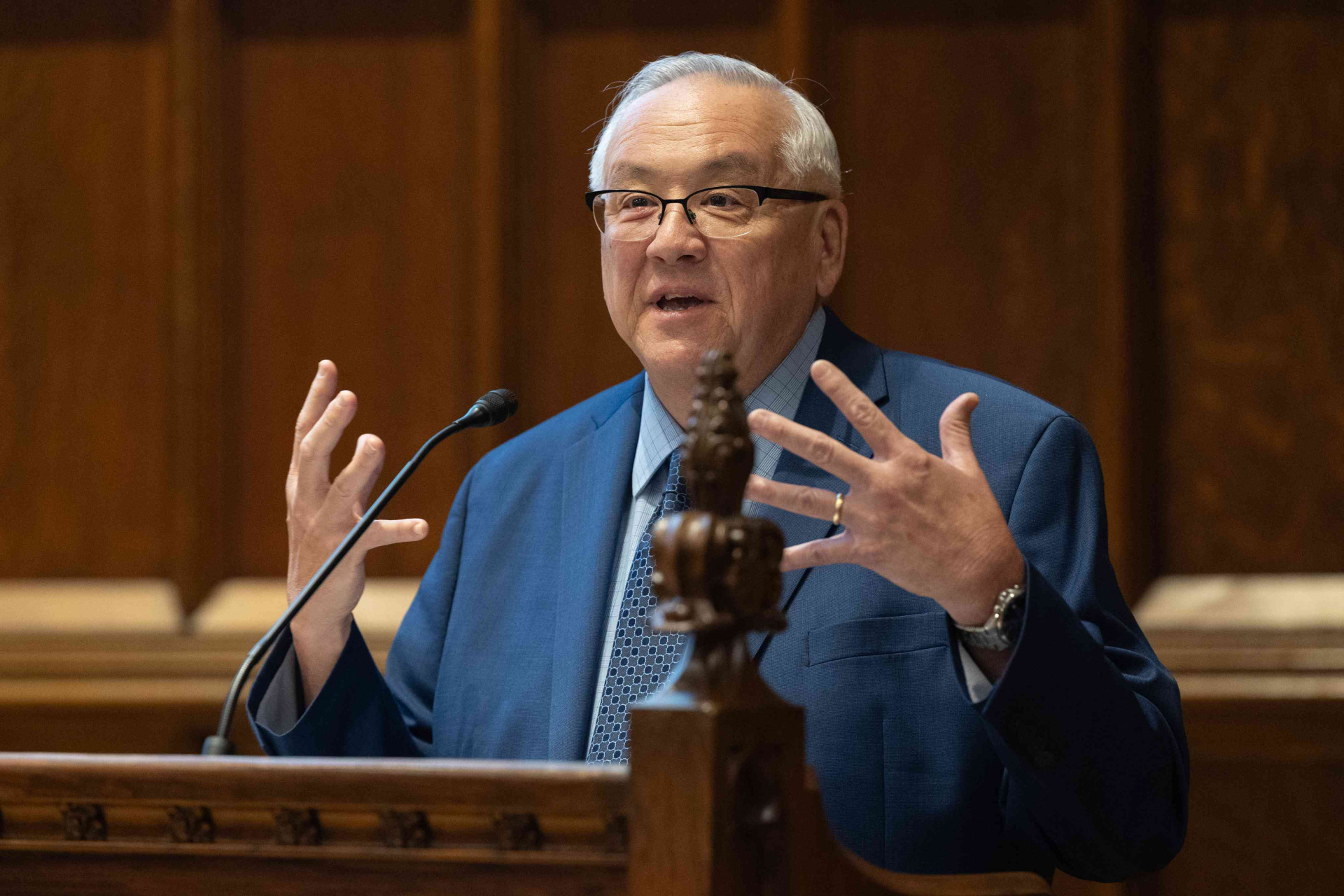A man with white hair wearing a navy blue suit and black glasses talks with his hands act a wooden lectern.