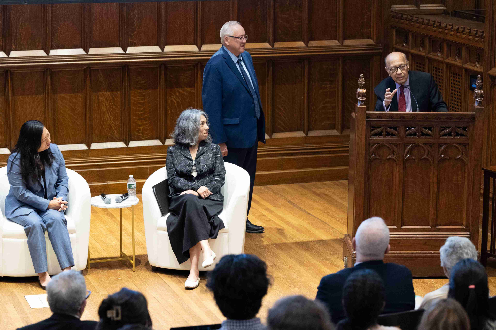 Two panelists sit in white chairs and listen to the speaker standing at a wooden lectern. A man stands to the left of the person speaking and smiles.