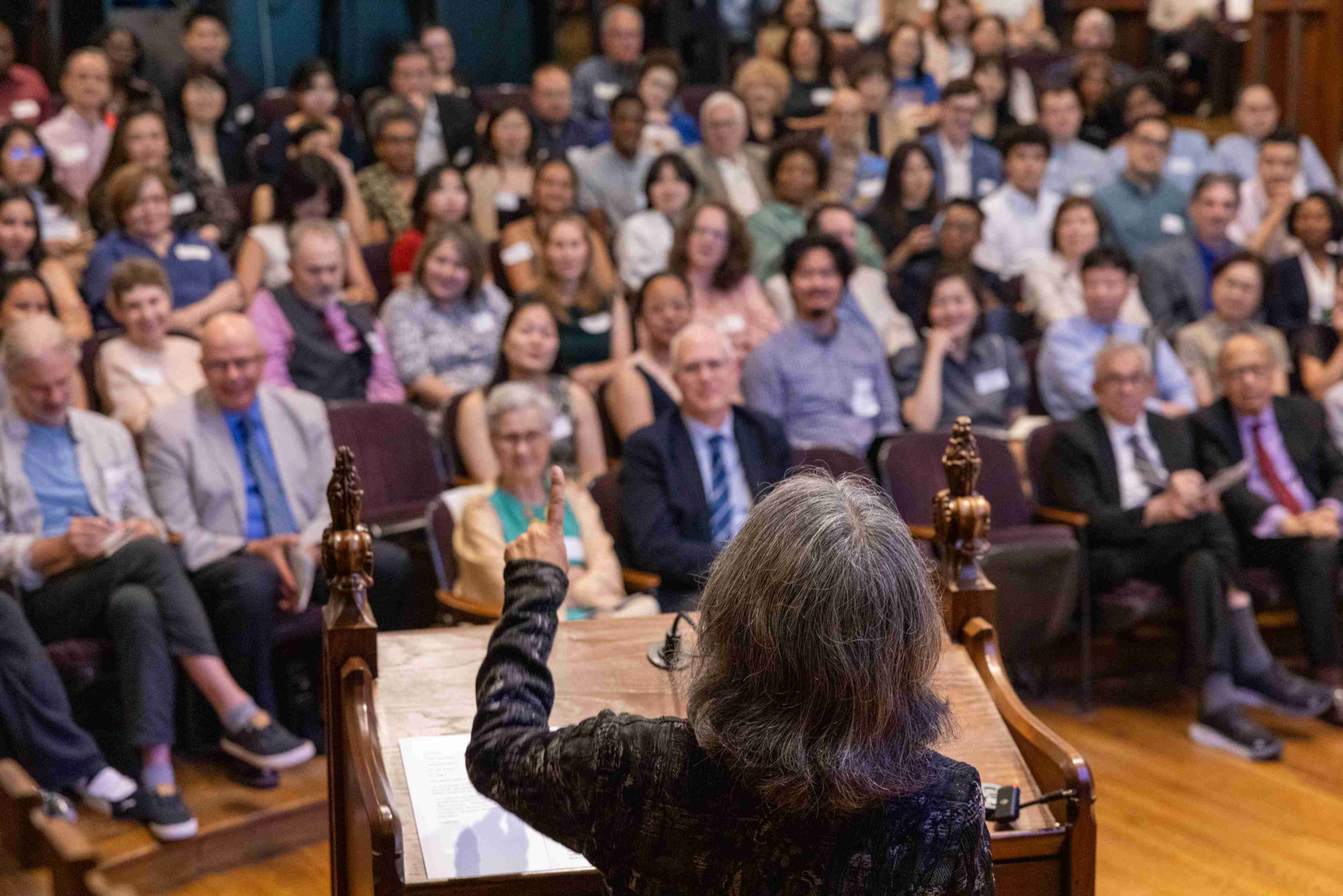 Image shot from behind: A person with gray hair wearing a black longsleeve holds up one finger as they speak from a lectern to a large audience of people.