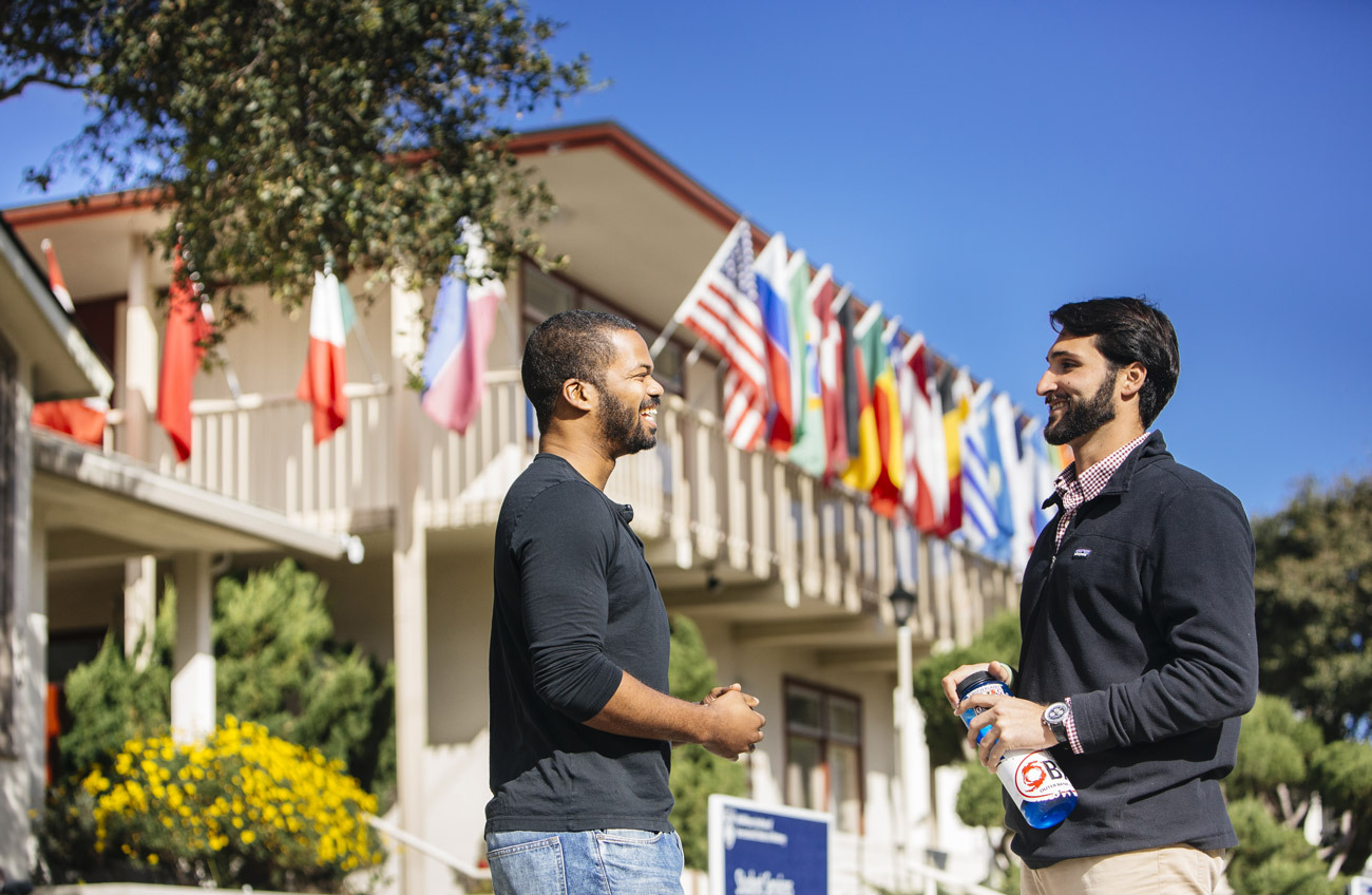 Two men wearing black longsleeve shirts smile as they converse outside a two-story building with various countries' flags hanging over the balcony.