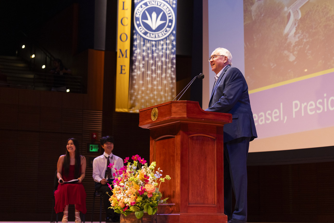 President Feasel wears a suit and speaks into the microphone from behind a wooden lectern. Two students are seated off to the left.