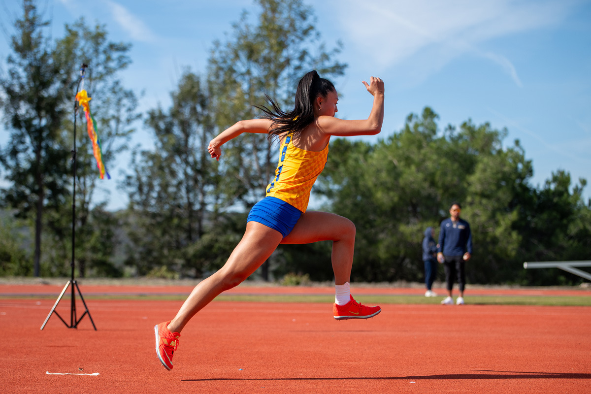 A female track athlete wearing a yellow tank top and blue shorts is mid-air on the orange track surface as she warms up.