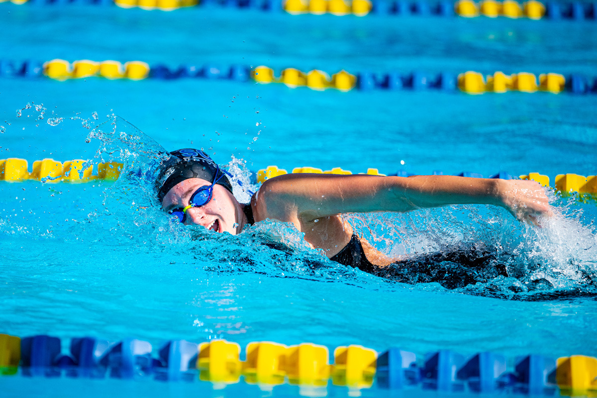 A swimmer wearing a black swim cap and blue goggles comes up for a breath as they swim freestyle in yellow and blue-lined lanes.