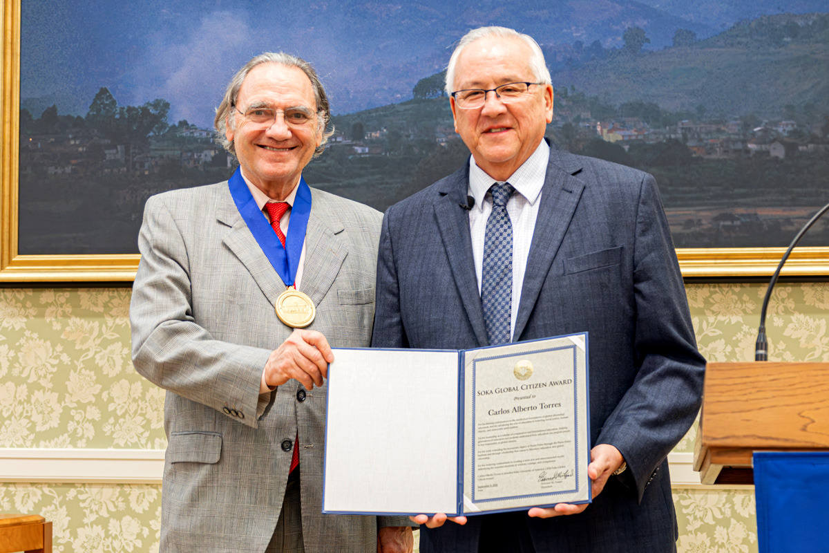 A man wearing a grey suit and a gold medal around his neck stands next to a man wearing a blue suit. They smile as they hold up an award.
