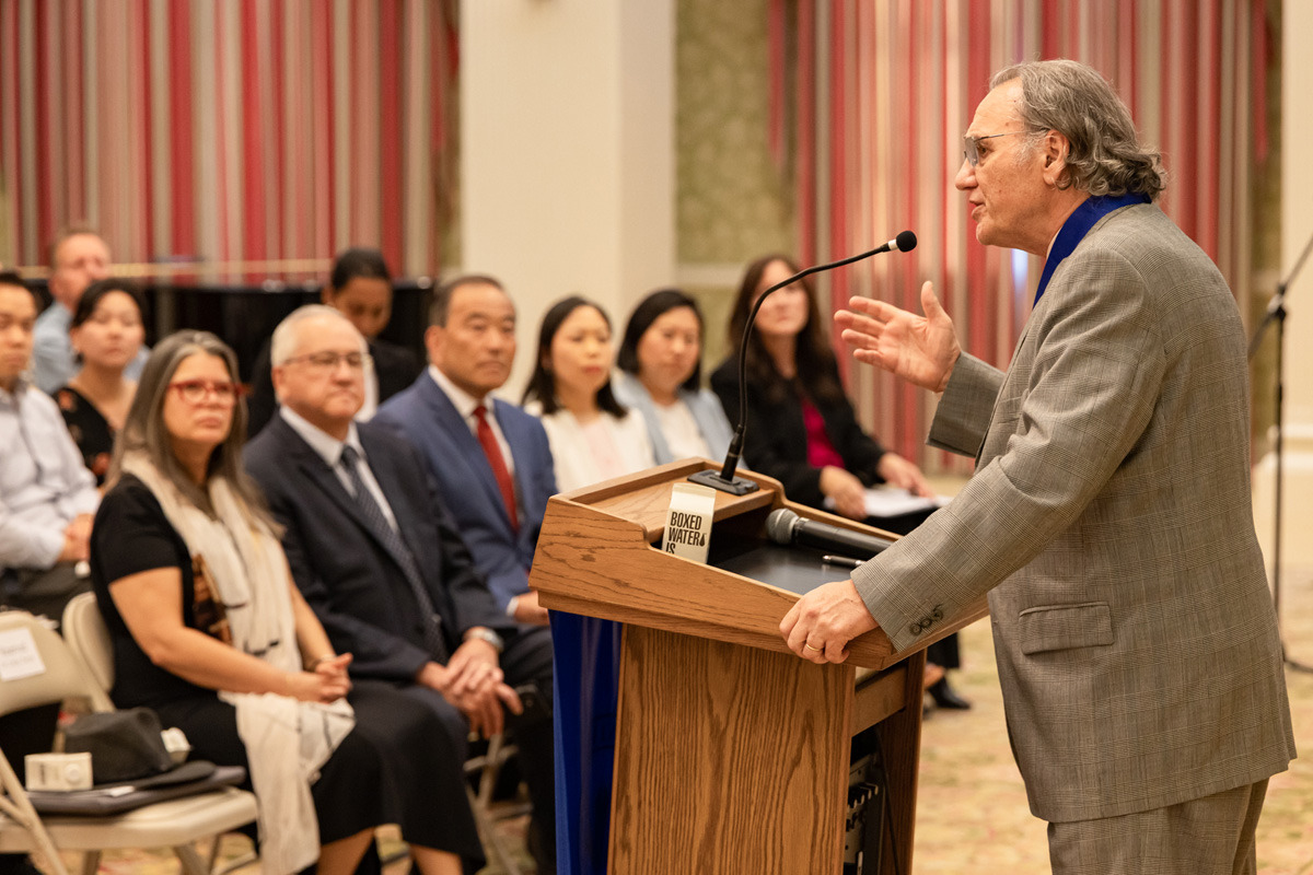 A man wearing a grey suit speaks into the microphone at a wooden lectern in front of a listening audience. There are multicolored pink and red curtains covering windows in the background.