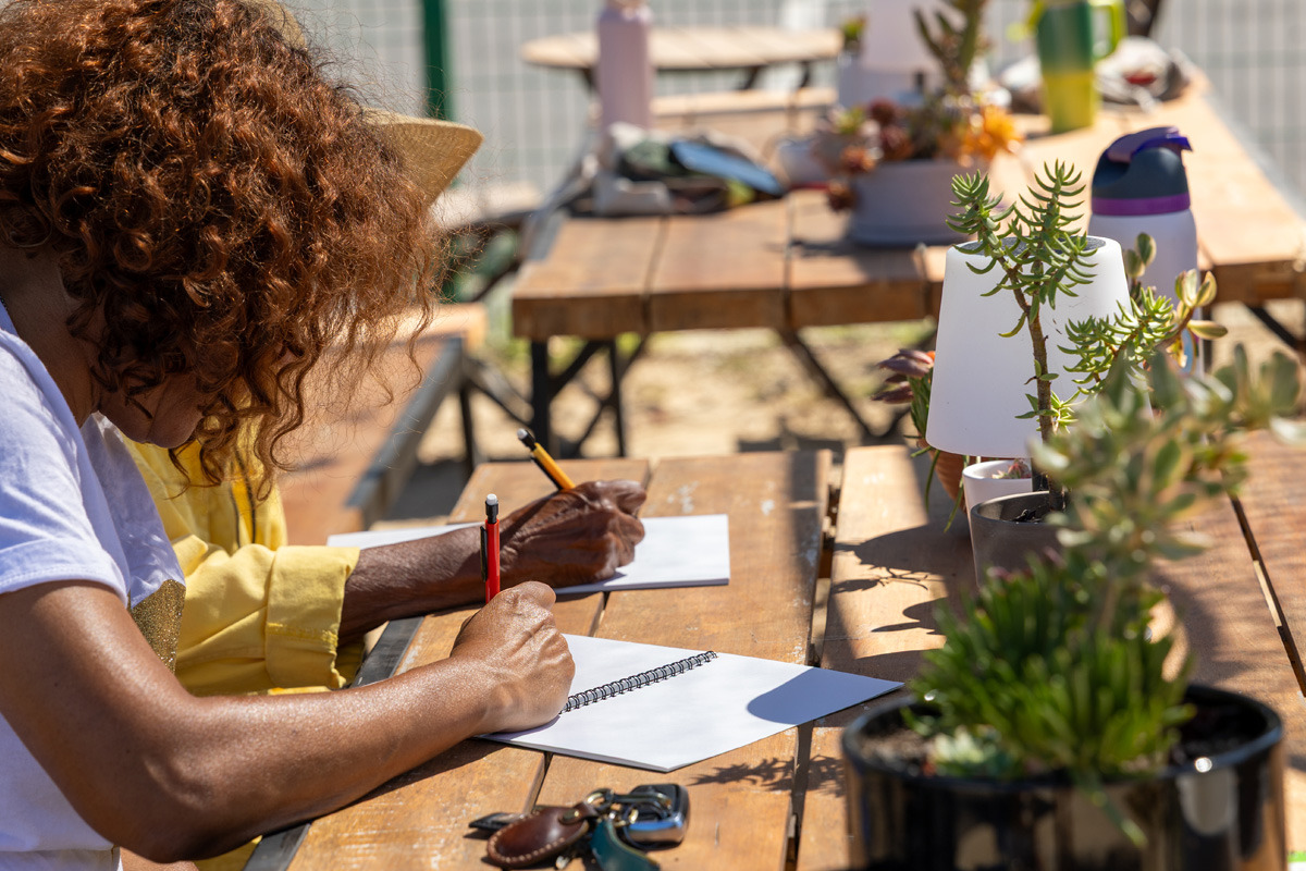 A woman with brown curly hair is writing in a notebook on a wooden table with plants on top of it.
