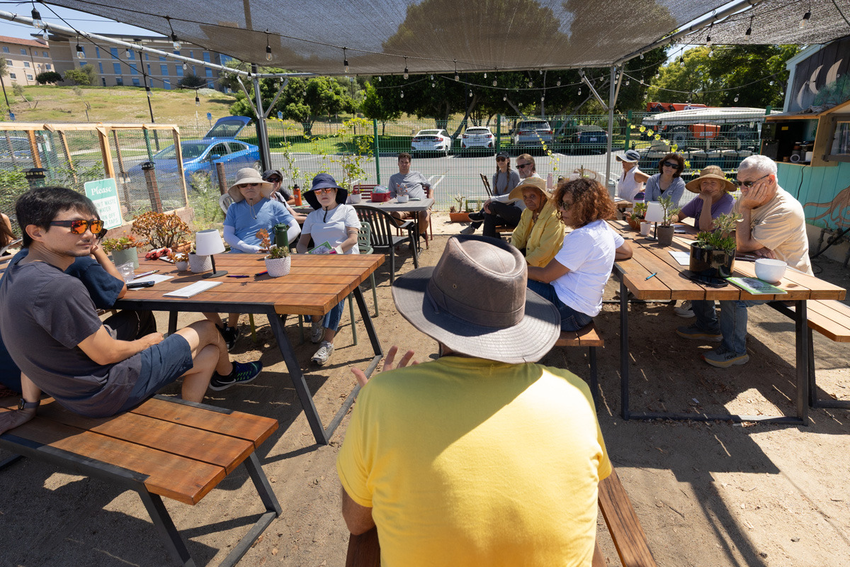A large group of people sit at wooden tables under a canopy in the garden on SUA's campus as they listen to a speaker wearing a yellow t-shirt and sunhat who has their back to us.