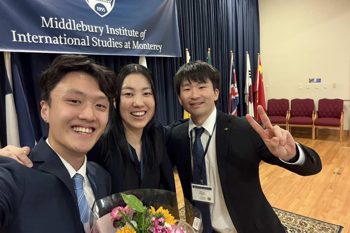 A woman stands in between two men in front of a navy banner that reads, "Middlebury Institute of International Studies at Monterey." They are smiling and holding a bouquet of flowers.