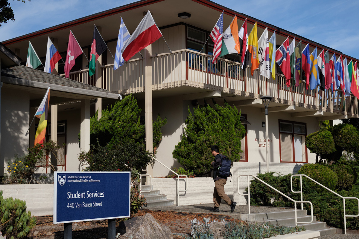 Exterior of a building on Middlebury's campus with various countries' flags hanging over the 2nd floor balcony. A person carrying a backpack is walking up the stairs towards the building.