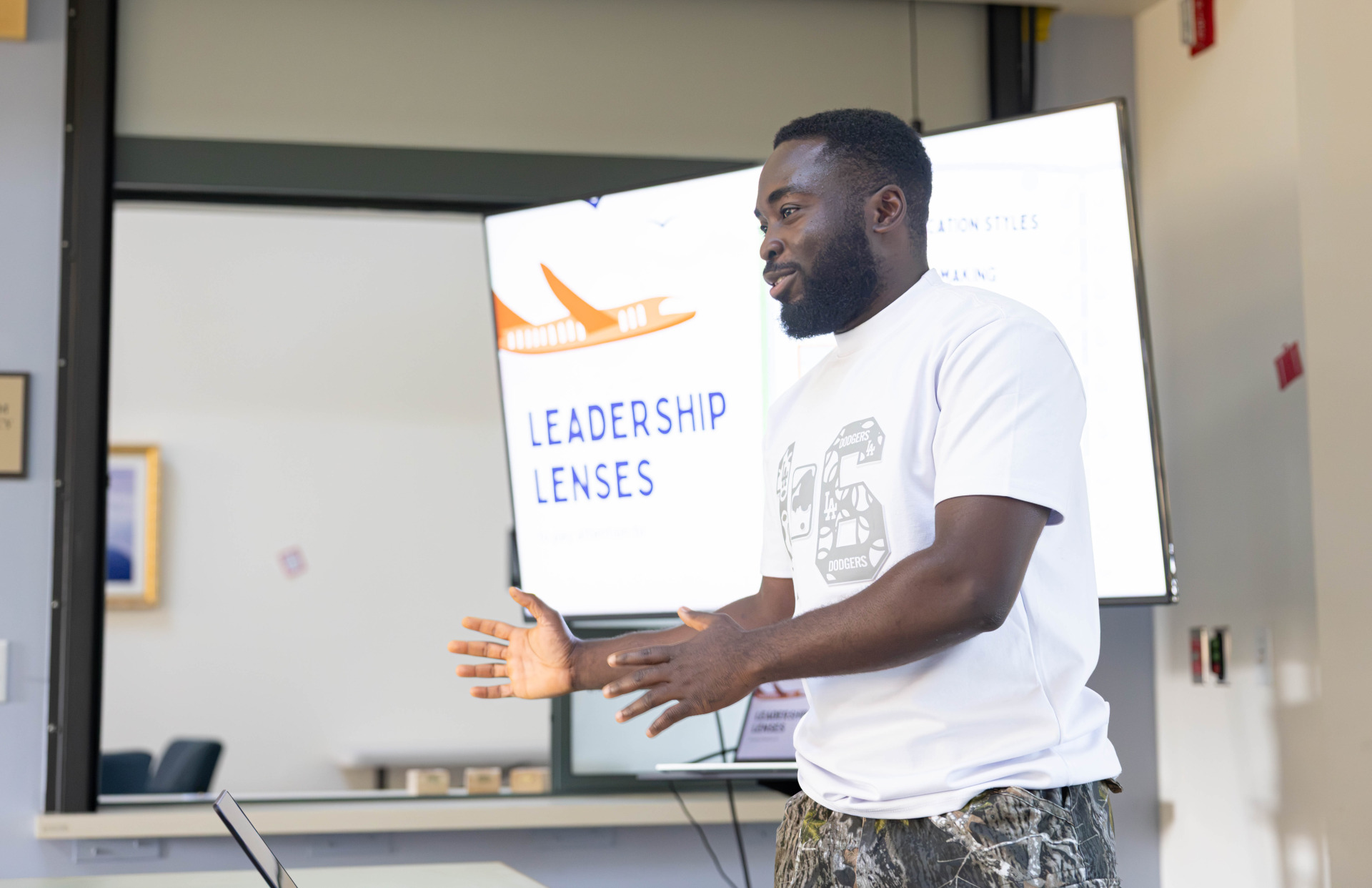 A man wearing a white t-shirt and camo pants gestures with his hands as he stands in front of a projector screen that reads, "Leadership Lenses."