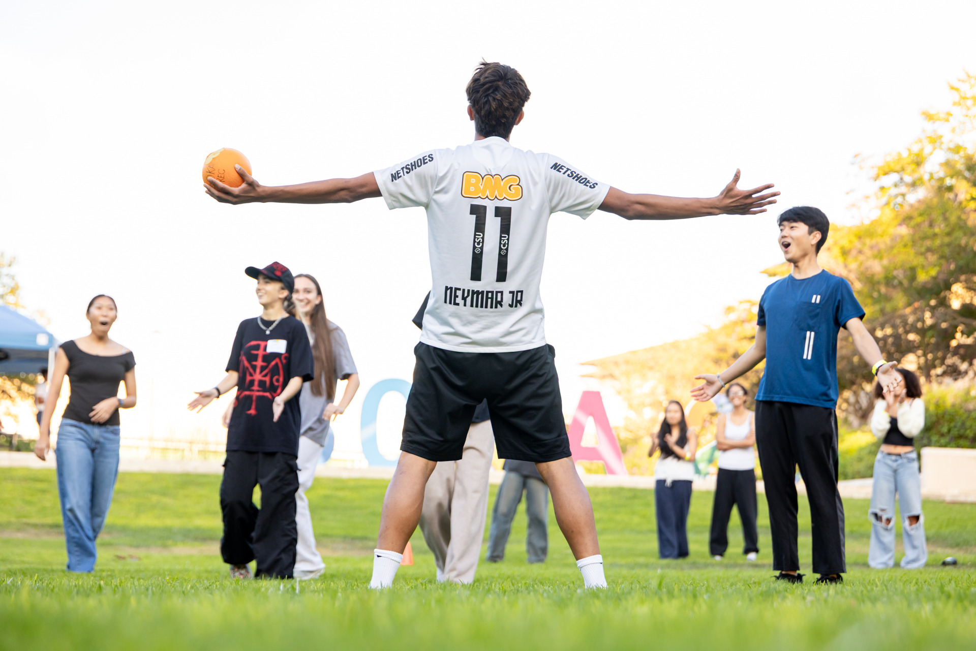 A student with short brown hair wearing a white Neymar jersey stands with his back to the camera and arms outstretched, holding a dodgeball in one hand as other dodgeball players look on.