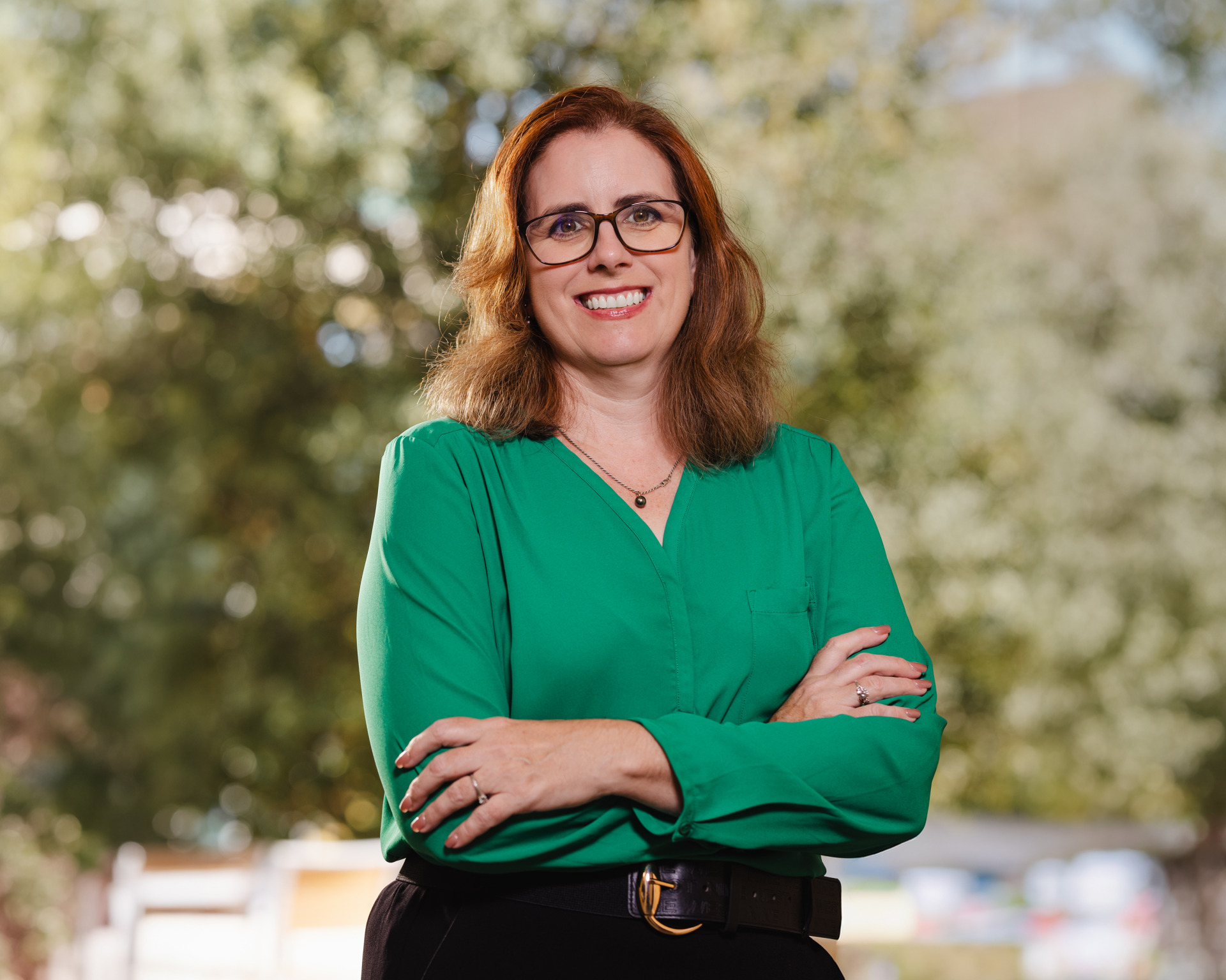 A woman with reddish-brown, shoulder-length hair is wearing a light green blouse, black pants, and black-rimmed glasses as she stands with her arms crossed and smiles. 
