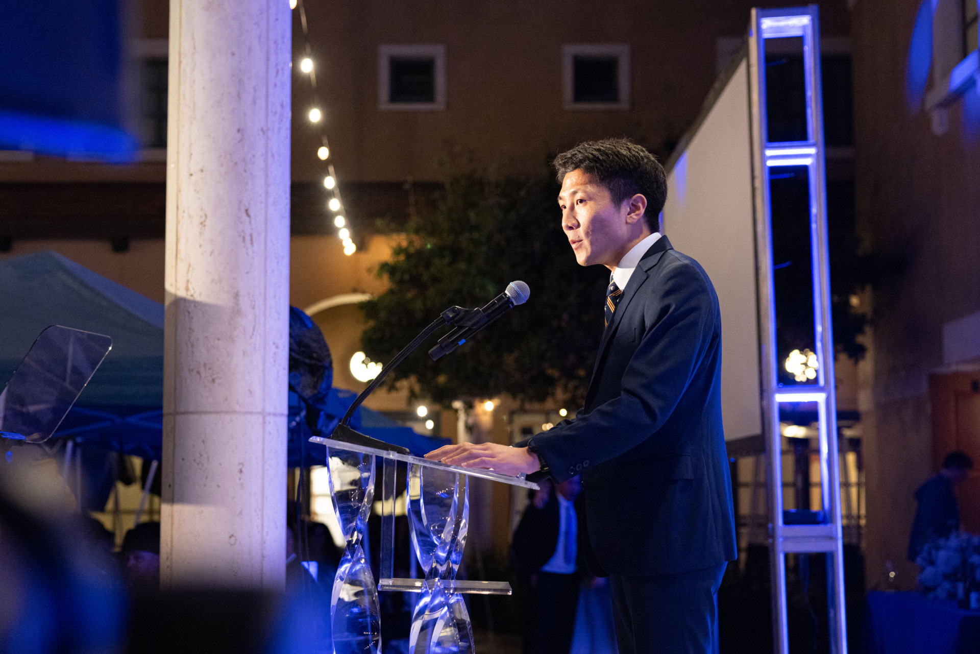 Side view of a man with short black hair wearing a black suit speaking into a microphone at a clear lectern with glass legs during the 2026 Peace Gala.