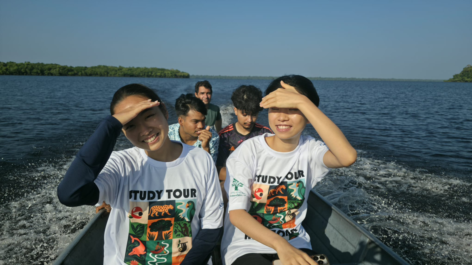 A group of five people are on a boat with green foliage far in the distance, the two people up front are using their hands to shield the sun from their eyes while smiling.