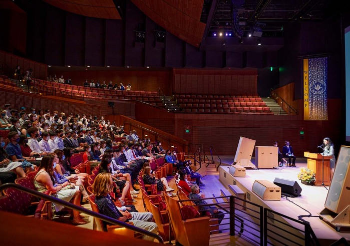 Person on concert hall stage speaks to students in audience