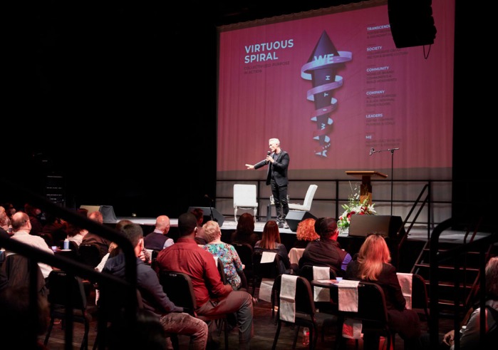 Simon Mainwaring holds a microphone and stands on stage in the Black Box Theatre on Soka's campus where he speaks to an audience about Rewiring Capitalism. There is a screen behind projecting some of his presentation points.