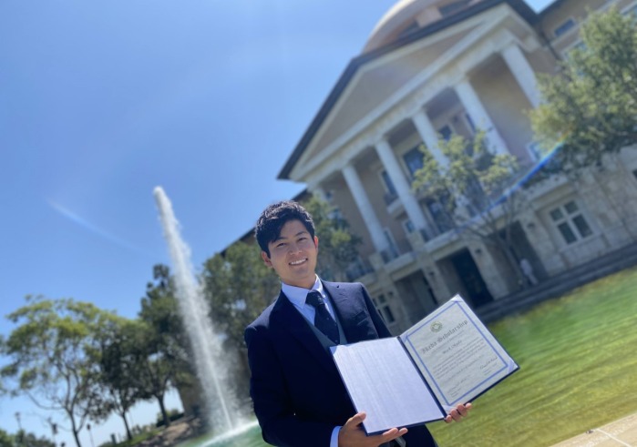 Mark Okuda, winner of the Frederick Douglass Global Fellowship, poses with his Ikeda Scholarship in front of Peace Lake and Founders Hall