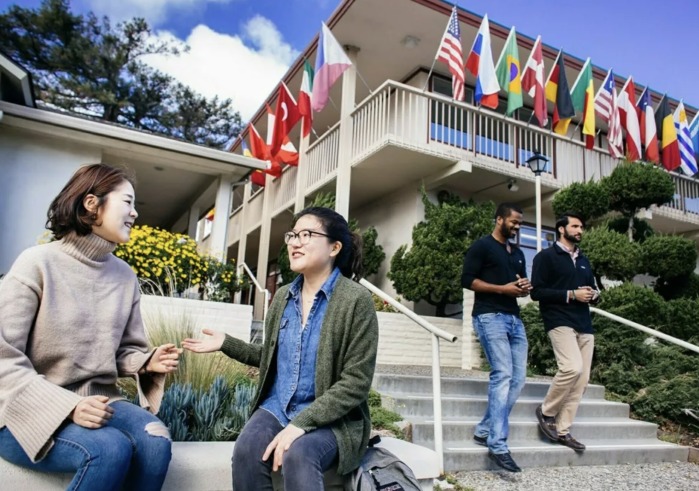 Two female students talk while sitting on a bench beneath a building displaying flags from various countries. Two men are walking down the steps to the side of the women talking.