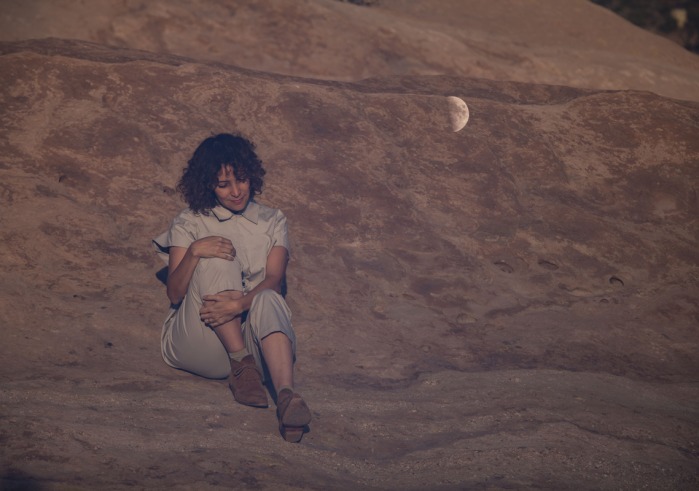 Gaby Moreno sits on a rock surface holding her knee to her chest. A half moon is on the rockface beside her.