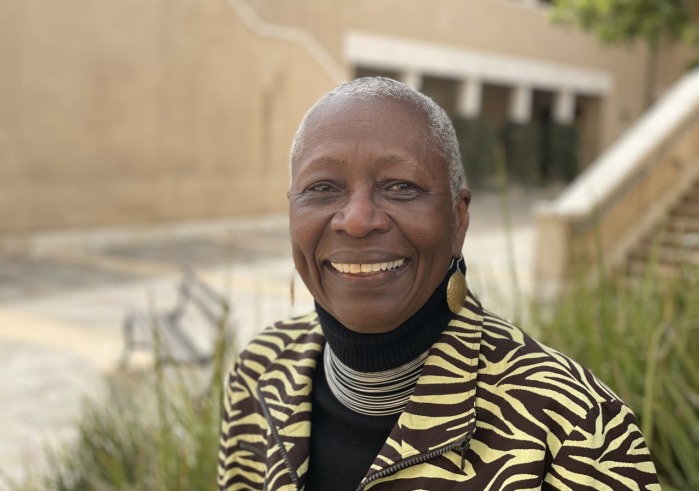 Professor Emeritus Gail Thomas smiles outside on Soka University's campus. She's wearing a black turtleneck with a zebra print zip up jacket and gold dangly earrings.