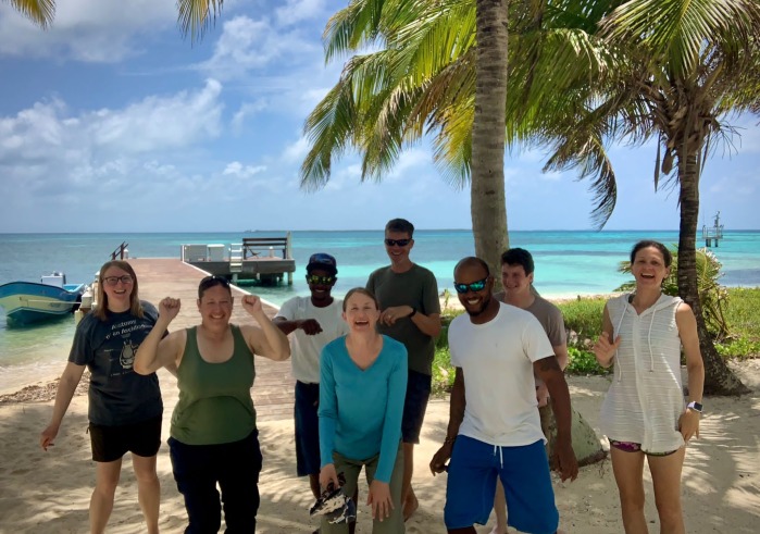 Eight members of a research team wearing summer attire smile at the camera in front of palm trees, a docked boat, and light blue water in Belize.