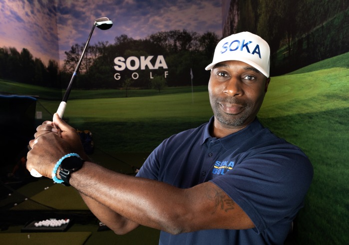Curtis Robinson, wearing a white Soka baseball hat, holds his golf club and poses in the Soka Golf training room on SUA's campus.